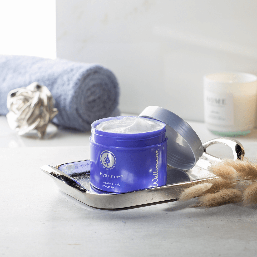 Hyaluron⁵ smoothing body mousse in a blue jar on a silver tray with a towel and candle in the background.