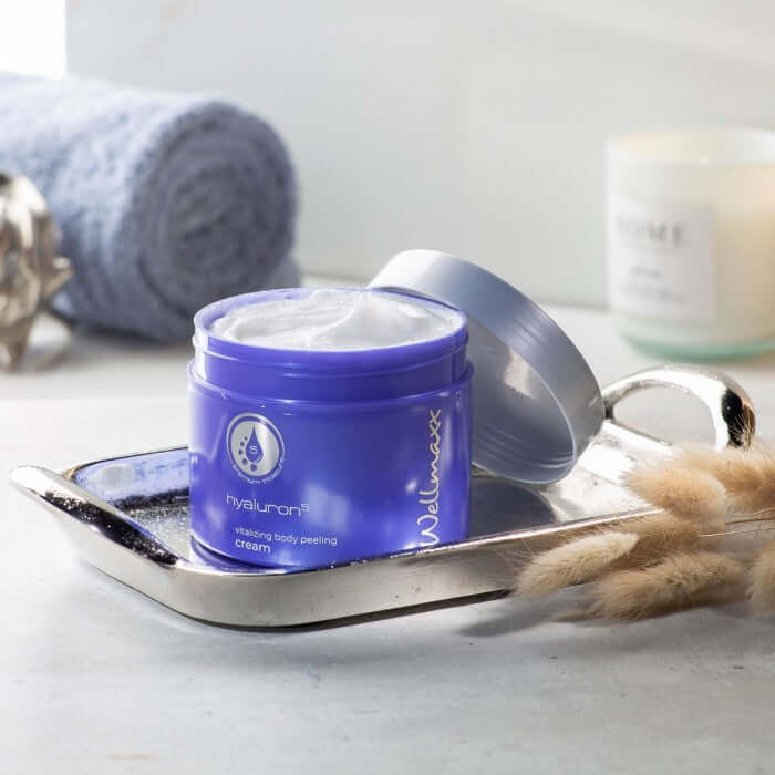 Hyaluron⁵ vitalizing body peeling cream in blue jar on a silver tray with towel, promoting smooth and revitalized skin.