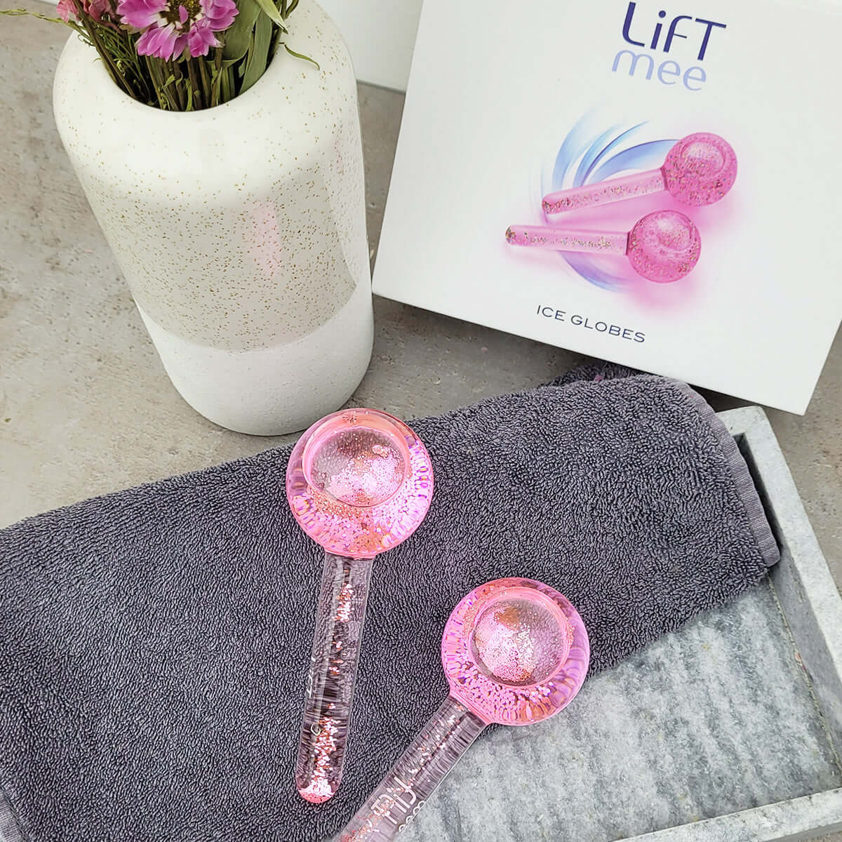 LiFTmee ICE GLOBES Duo Set in pink, placed on a grey towel with a vase and box, ideal for facial massage and skincare.