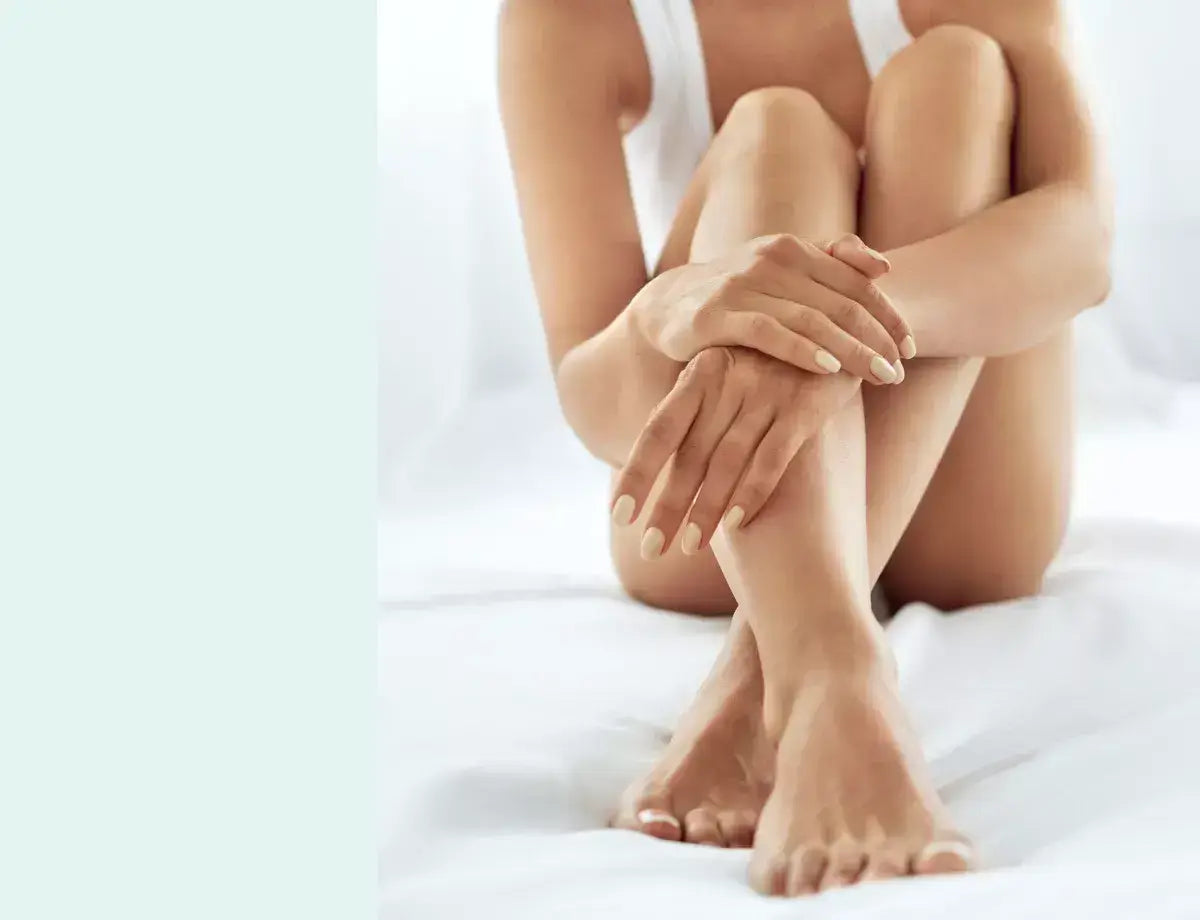A close-up of smooth legs and hands resting elegantly on a white bed, showcasing natural nail polish and a serene atmosphere.