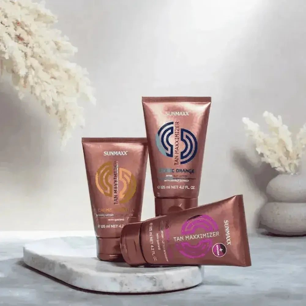 Drei Tuben Tan Maxximizer Creme Caramel Bräunungslotion auf Marmorplatte mit zarter Deko im Hintergrund